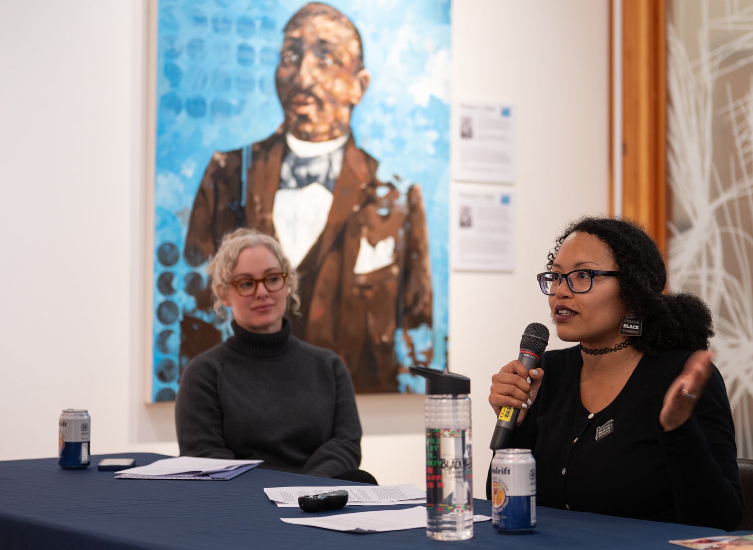 A Black woman speaks into a microphone as a white woman looks on.