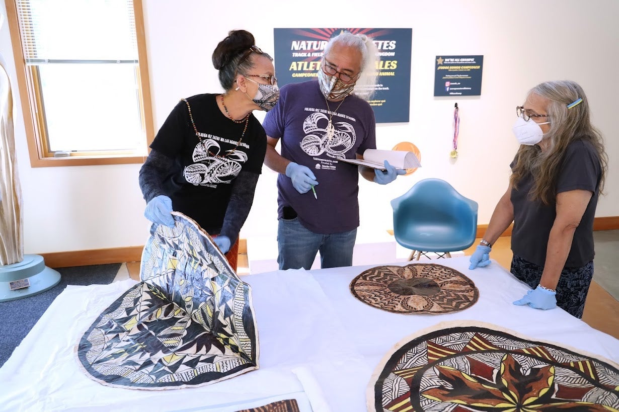 Two Samoan tapa researchers investigate a tapa cloth as MNCH collections director looks on