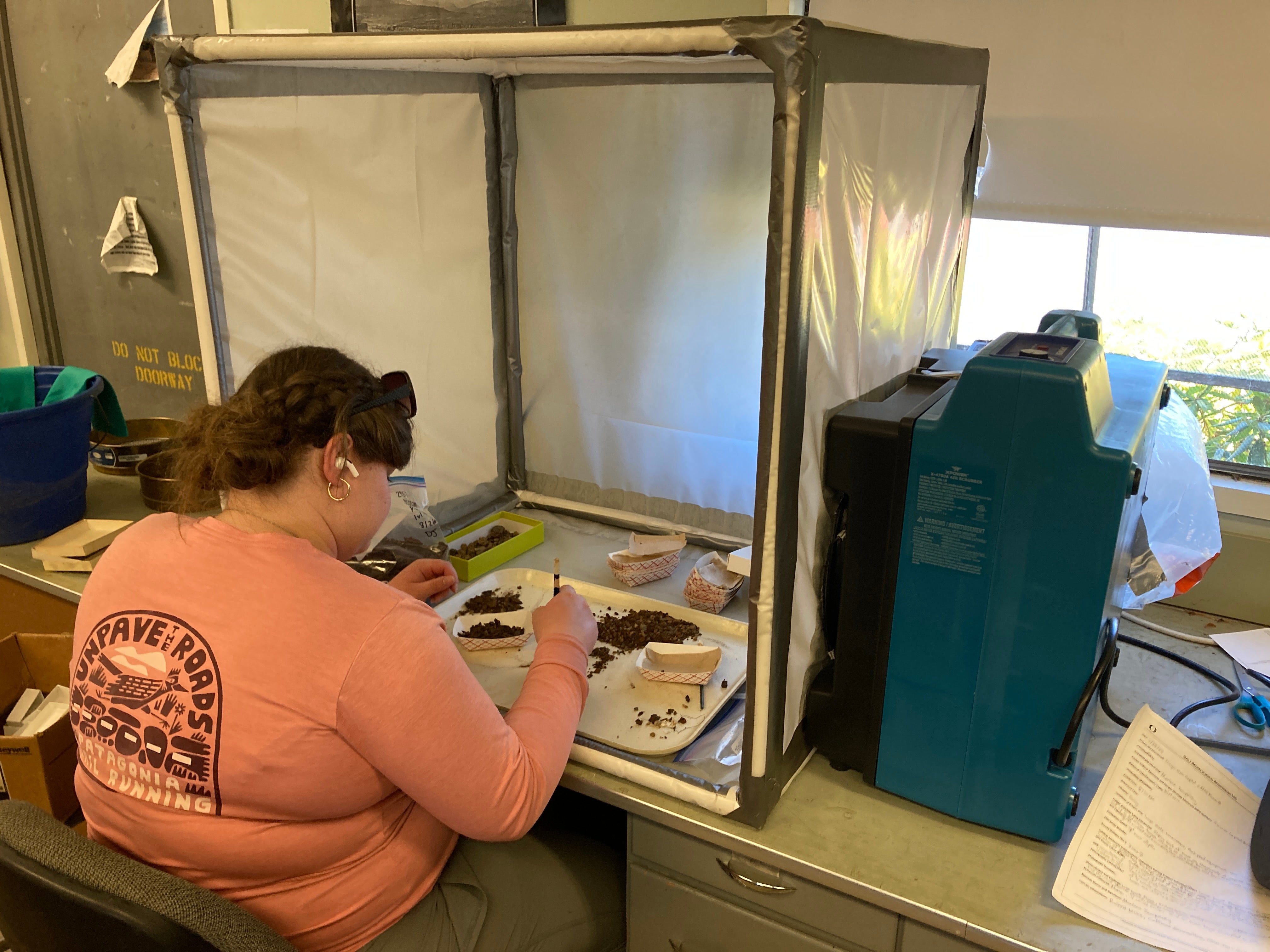 An archaeologist sits with their back to the camera, surrounded by a plastic "hood" with a large fan. They are working on small bits of metal.