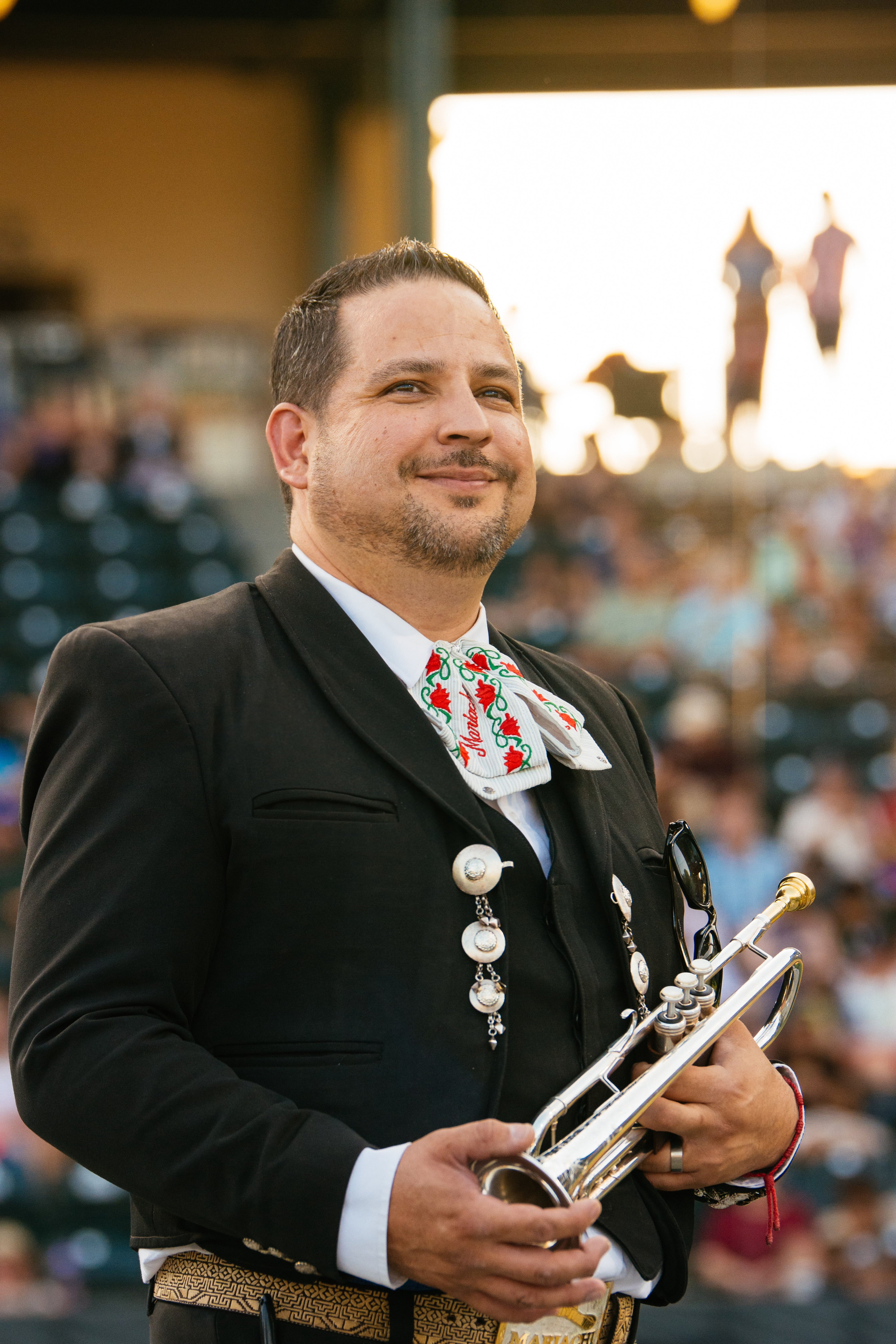 Arturo stands posing in a stadium, with his trumpet and wearing traditional mariachi regalia.