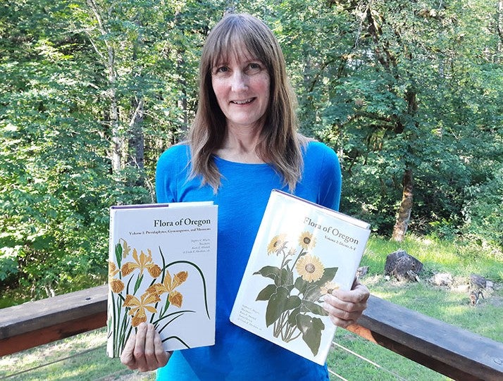A white woman in a blue shirt is holding two books about Oregon flora. 