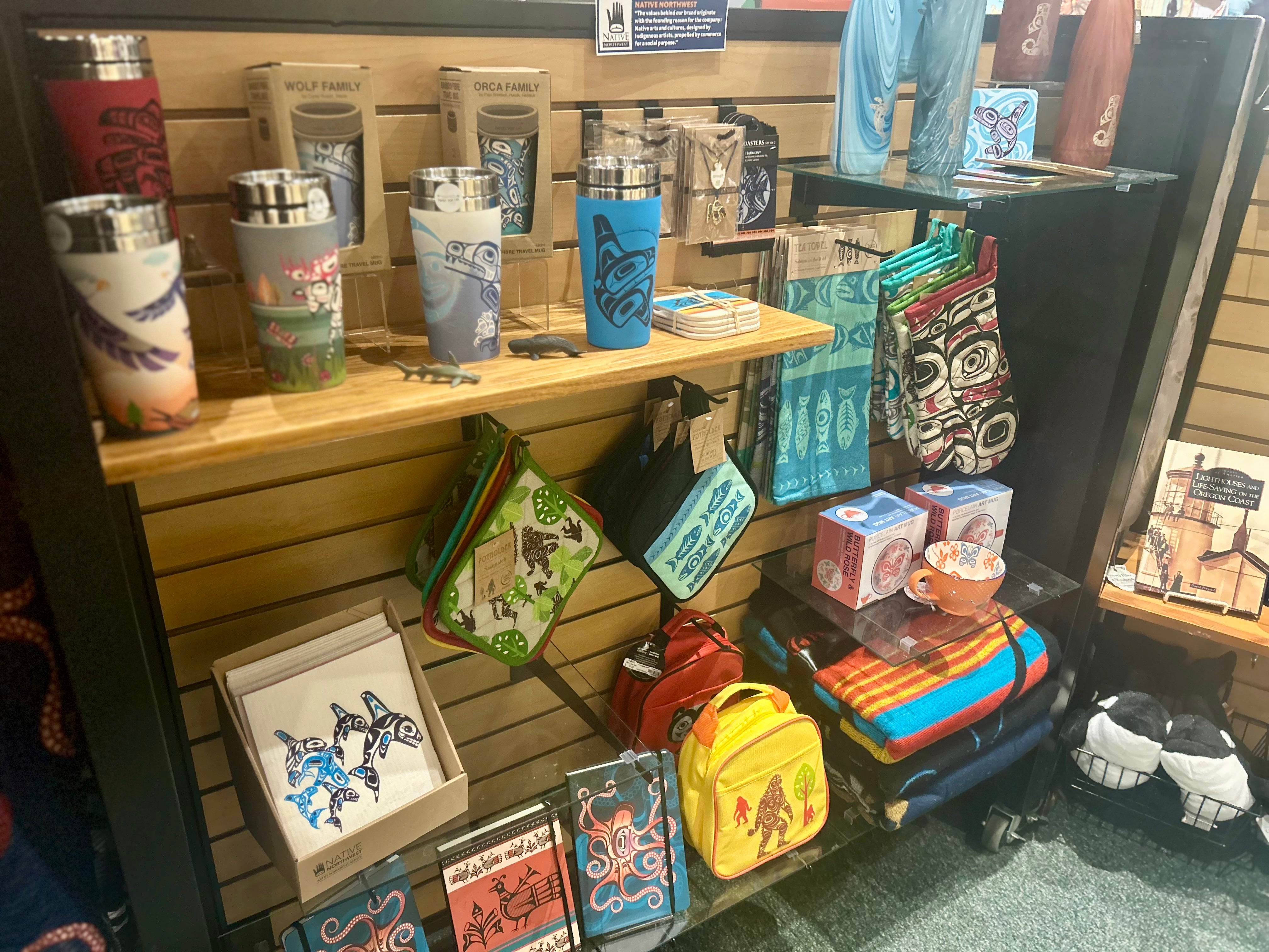 Coasters, a travel mug, a backpack, and a tea towel, all with Pacific Northwest Native motifs. The travel mug features a moose and the backpack features Bigfoot.