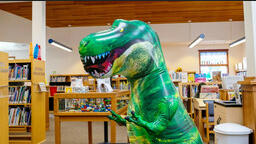 A green inflatable T Rex dinosaur stands in a library children's section.