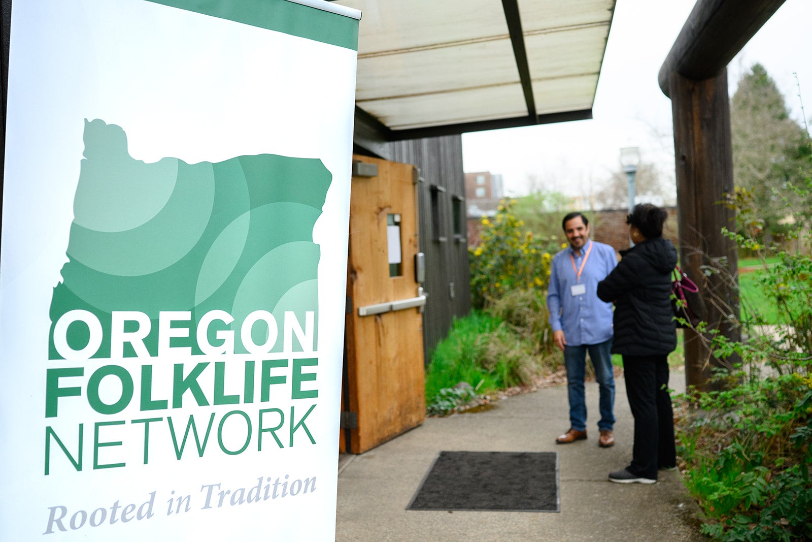 Two people stand to the right of the Oregon Folklife Network Banner 