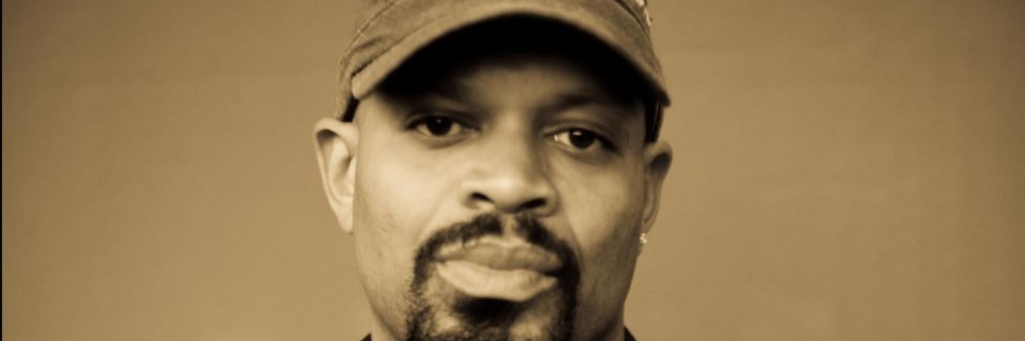 Sepia toned headshot of Mic Crenshaw, emcee. Mic is a black man with a goatee, wearing a denim jacket and baseball cap.