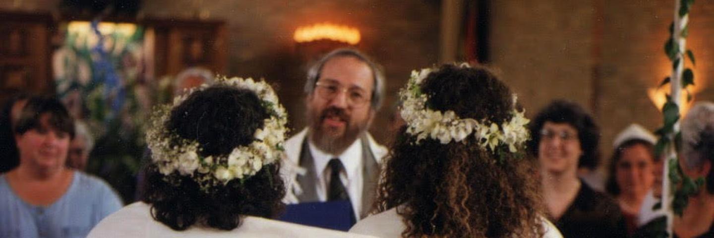 The backs of two women in white, with one shawl across both of their shoulders, two flower crowns, facing a rabbi