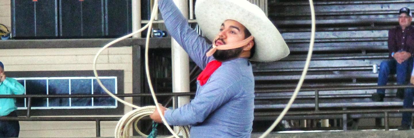 Josue Mendoza riding a horse and performing rope skills. He is wearing a white hat, blue shirt, and leather chaps.