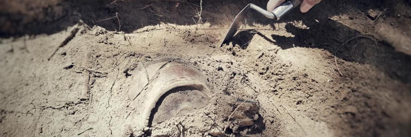 A close up on a trowel scraping dirt away from a half-buried ceramic object
