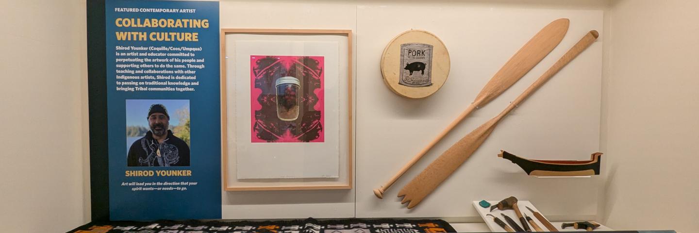 An exhibit case. A panel is on the left with visible text reading "Featured Contemporary Artist: Collaborating with Culture." Inside the case is a framed art piece featuring berries in a mason jar (evoking Andy Warhol's pop art), carved canoe paddles, and a colorful woven blanket. 