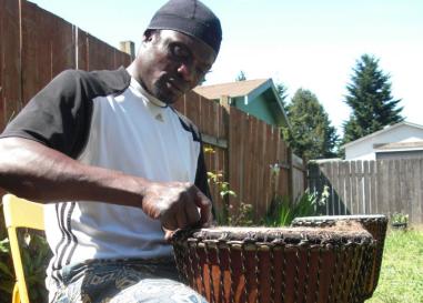 black man in white shirt and patterned pants working on a drum