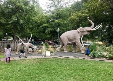 Three children walking like mammoths in front of an outdoor mammoth sculpture