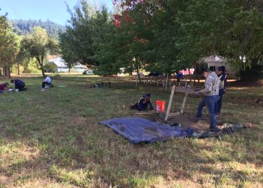 Archaeologists in a grassy field. Some are digging, some are measuring, and some crew shaker screens. 