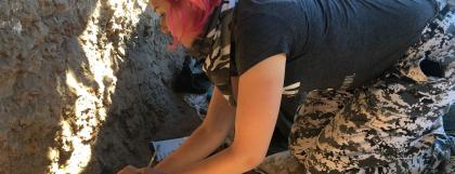 Woman kneeling on ground, excivating at the field school site.