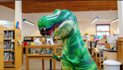A green inflatable T Rex dinosaur stands in a library children's section.