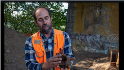 A white man with a high-viz vest holds a broken and dirt-encrusted piece of a wine bottle under a bridge.