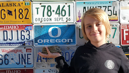 a young boy with long blond hair holds a mammoth tooth against a wall of license plates