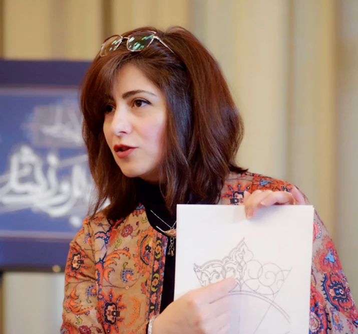 Photo of Marjan Anvari, a Persian woman with brown hair and glasses pushed up on her head. She is pointing at a paper with calligraphy on it.