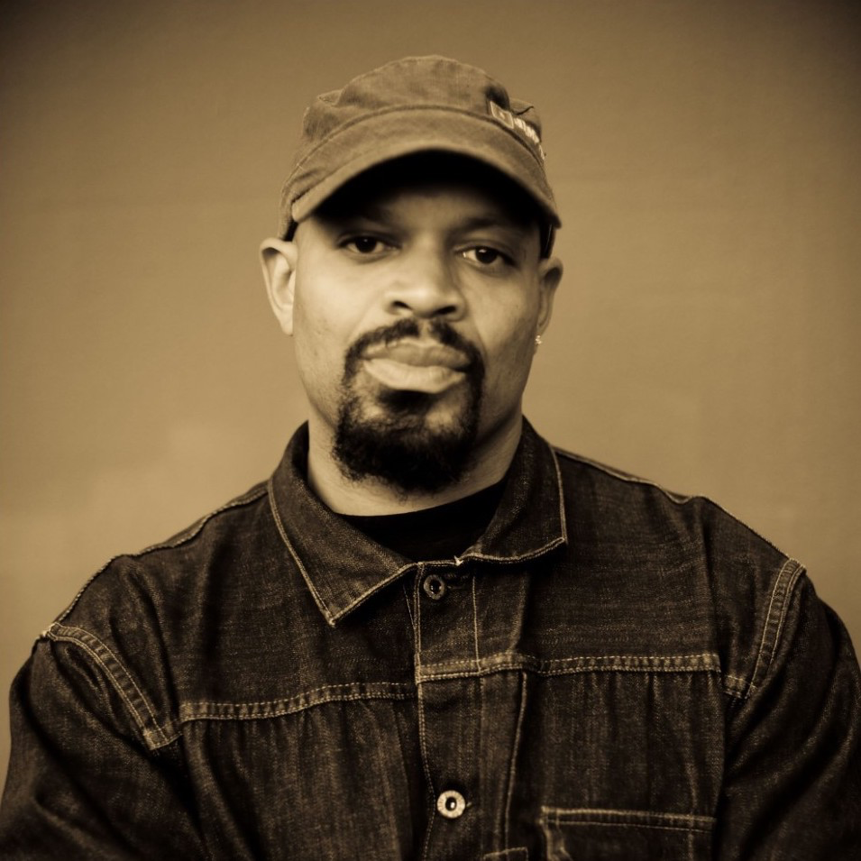 Sepia-toned headshot of Mic Crenshaw, a Black man with a goatee, a dark denim jacket, and a hat.