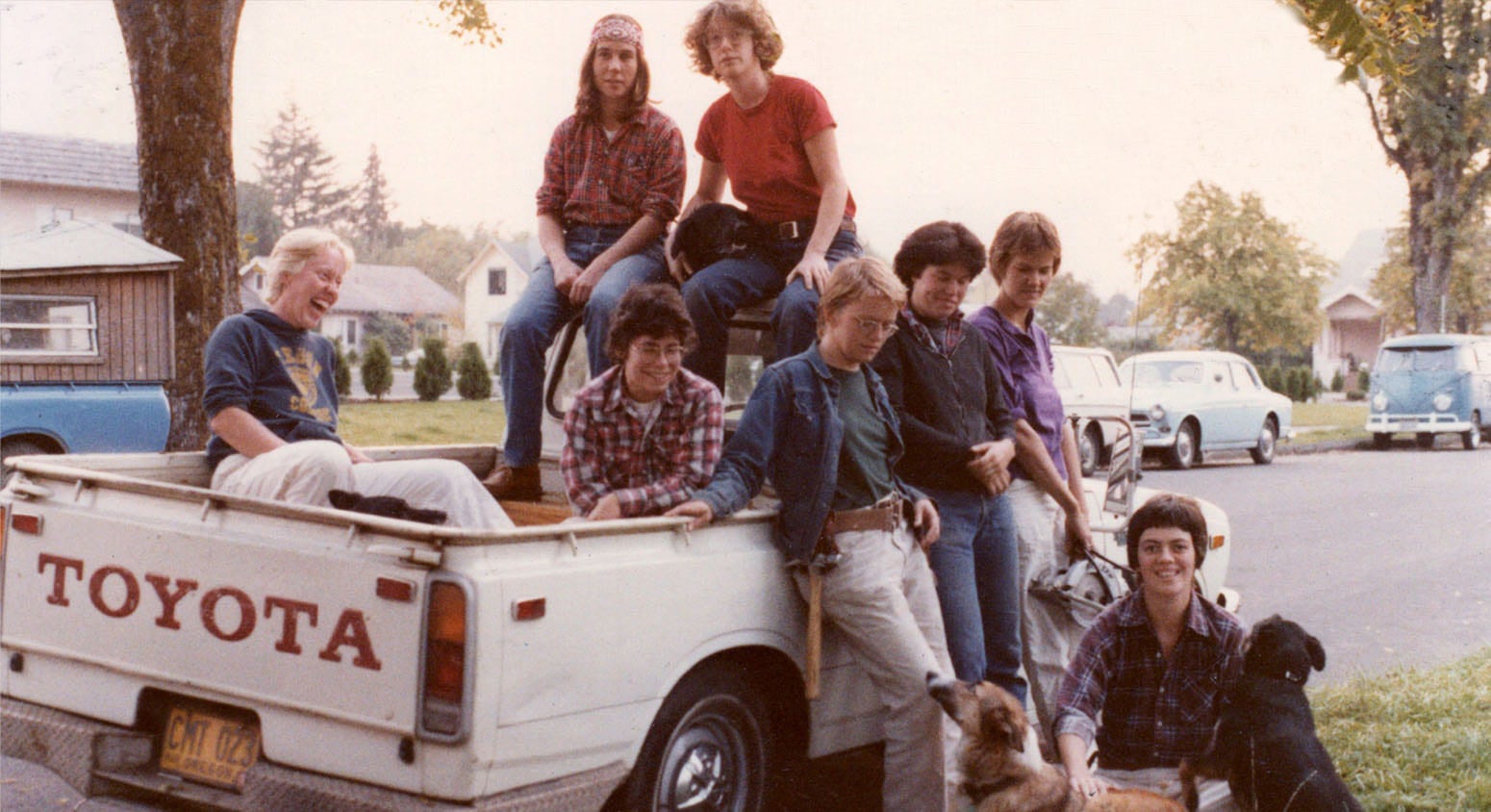 Several white women with short hair identifying as lesbians lean against a white Toyota pickup truck. There are four dogs in the image.