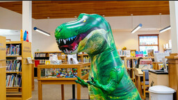 A green inflatable T Rex dinosaur stands in a library children's section. 