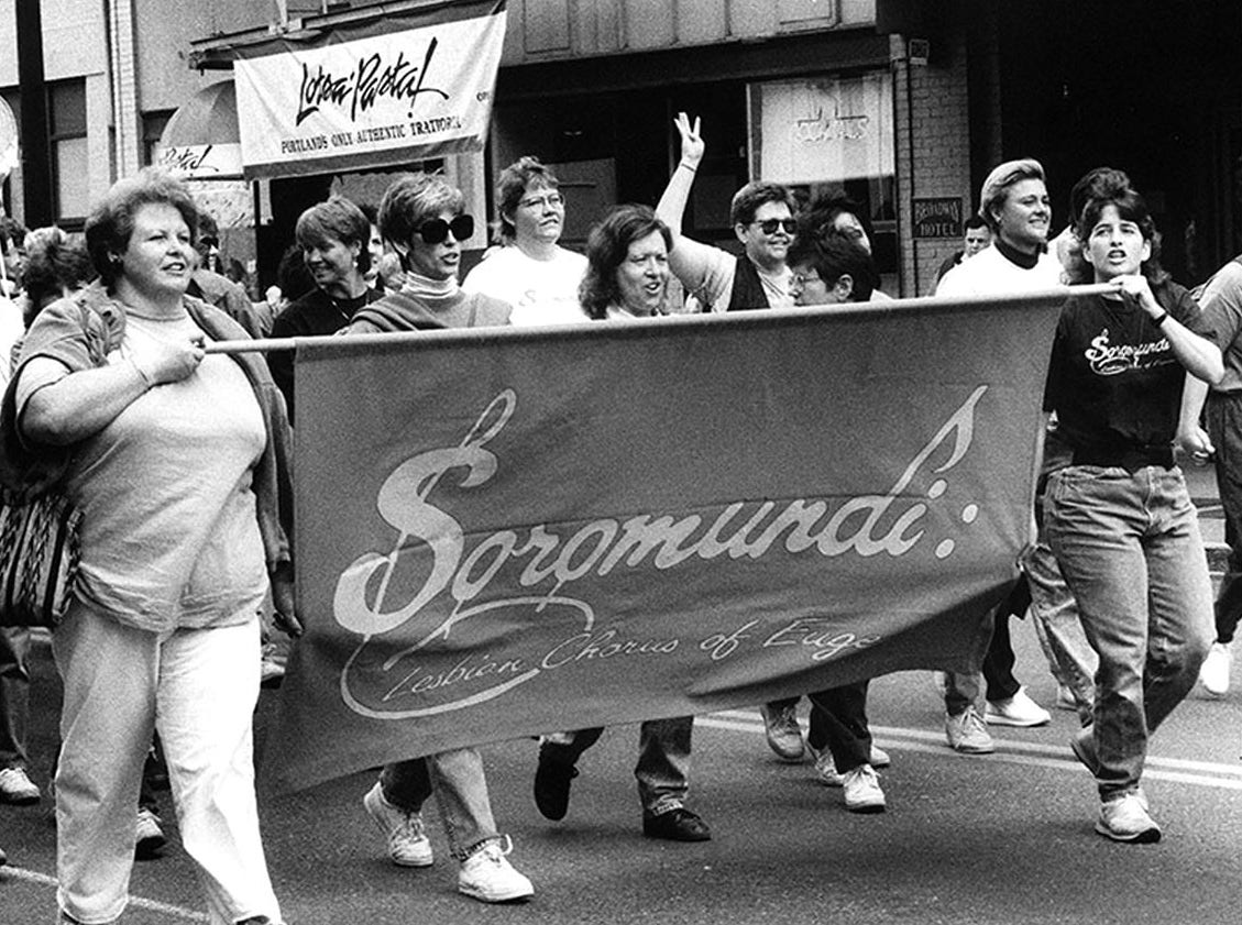 A black and white photo of women holding a banner marching in a parade. The banner reads Soromundi: Lesbian Chorus of Eugene