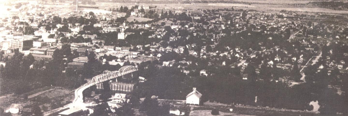 Black and white aerial photo from 1923 showing the Van Buren bridge, a truss bridge, and the city of Corvallis and surrounding mountains.