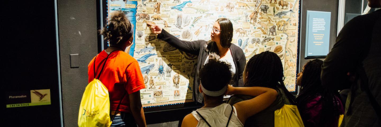 A young woman with long, black hair points to a fossil map of Oregon while visiting high school students listen and learn about natural history