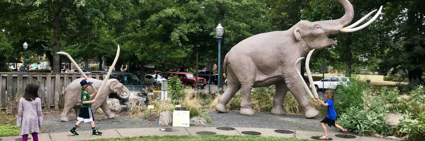 Three children walking like mammoths in front of an outdoor mammoth sculpture