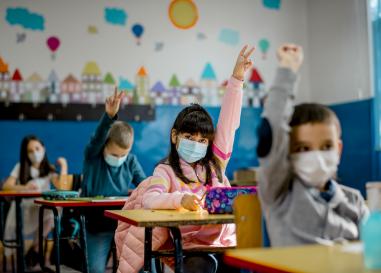 Students wearing masks in a classroom, many of whom are raising their hands