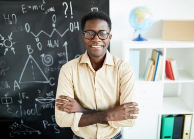 A teacher smiles at his class with a chalkboard and books in the background.
