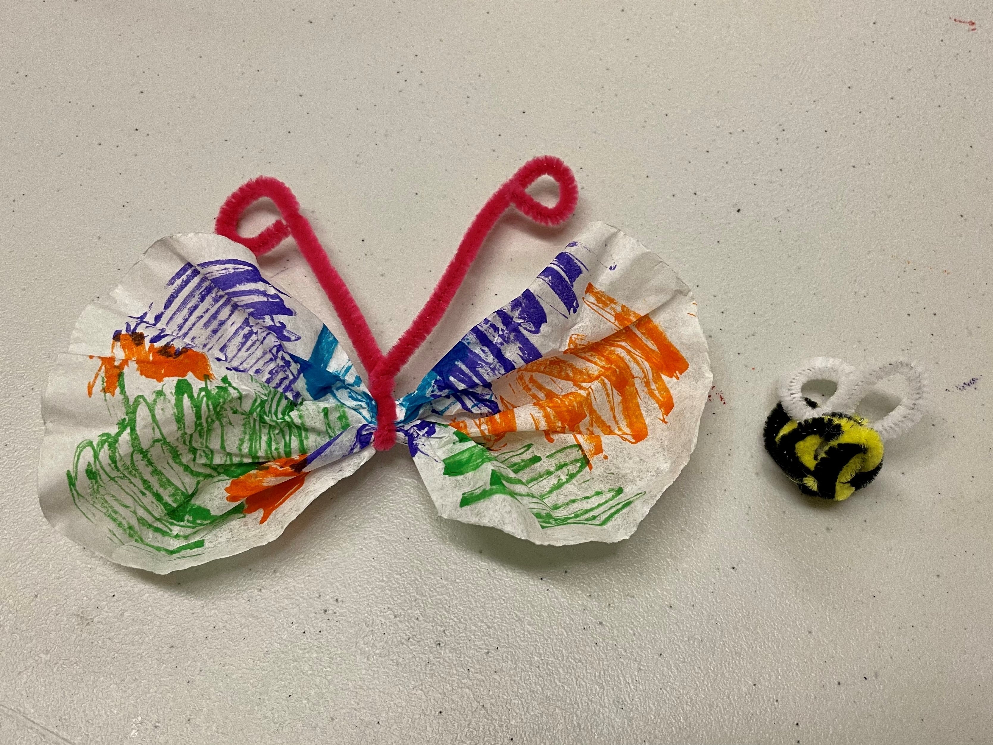 A coffee filter that's been colored and scrunched in the center to look like a butterfly, next to a bee made from black and yellow pipe cleaners