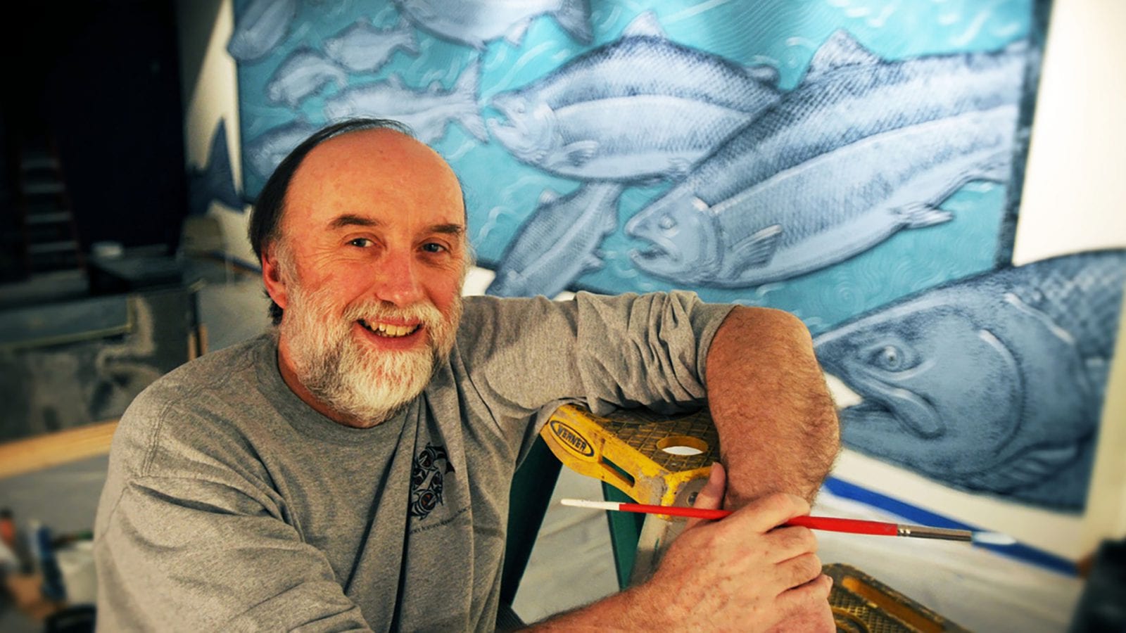 Ray Troll smiling and holding a paintbrush. One of his fish murals is visible in the background; it's multiple shades of blue and features Troll's signature quirky style. 