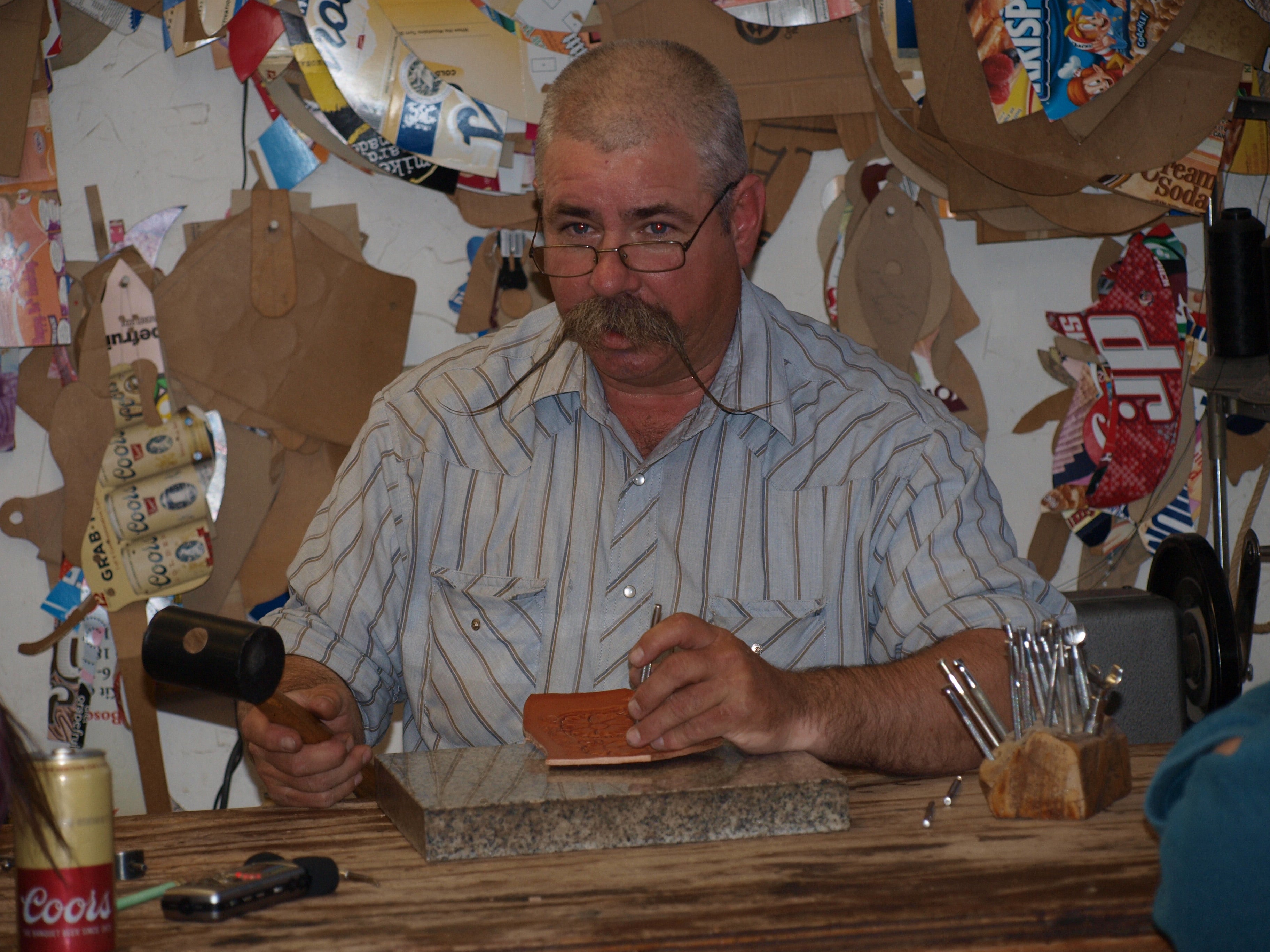 Image of Steve McKay, a white man with short gray hair, wire rim glasses, and an enormous mustache. He's holding a mallet and tapping a scrap of leather. The wall behind him is hung with cardboard saddle patterns.